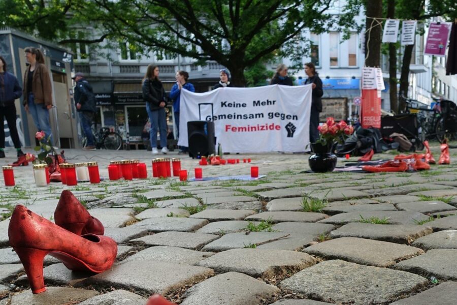 Bild von einer Kundgebung des Anti-Feminizid-Netzwerks auf dem Alma-Wartenberg-Platz in Hamburg. Im Vordergrund steht ein rotes Paar Absatzschuhe, im Hintergrund sind Grabkerzen zu sehen.