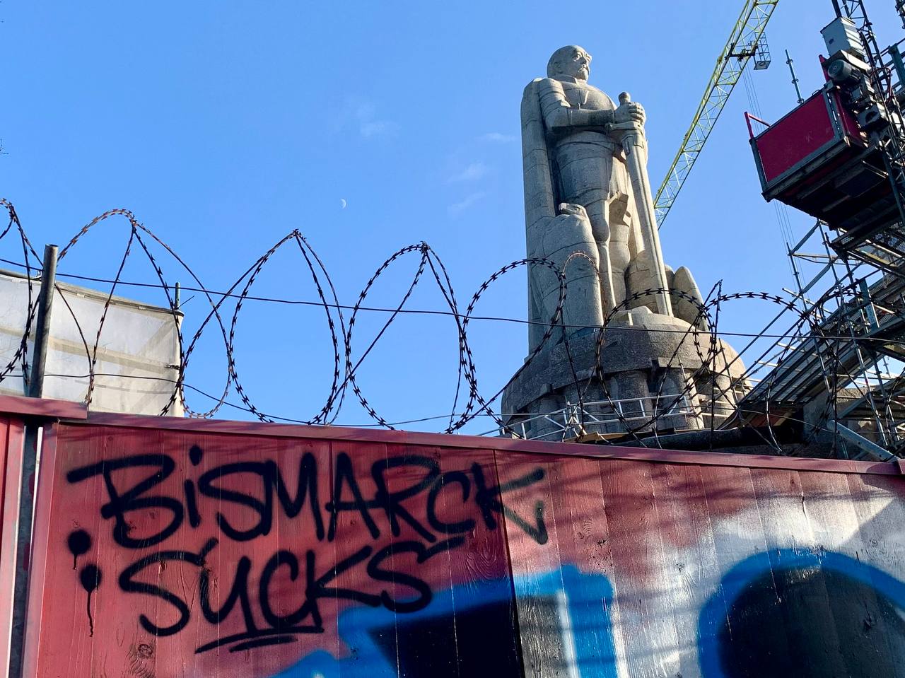 Eine Mauer mit Stacheldraht auf der "Bismak Sucks!" geschrieben steht. Im Hintergrund ist die Hamburger Bismarkstatue zu sehen.